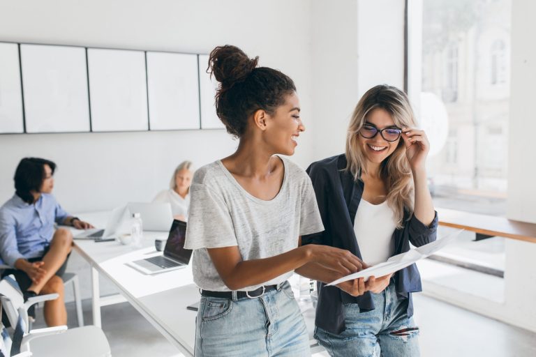 Young female executive explaines new strategy to blonde employee in glasses and smiling. Indoor portrait of multicultural collective working on project in office and using laptop..